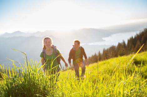 2 Personen wandern im hohen Gras, im Hintergrund sind Berge und ein See zu erkennen