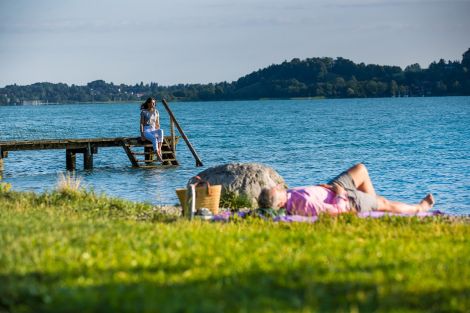 Eine Person liegt auf dem Gras am Seeufer, im Hintergrund eine Person auf einem Steg
